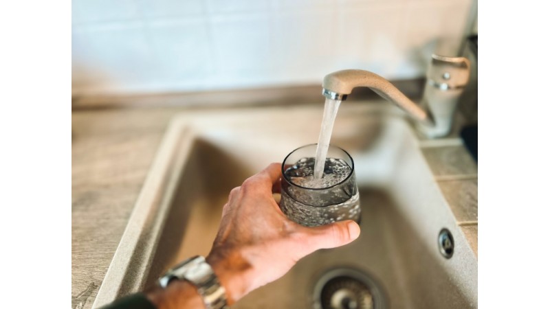 Męska ręka trzymająca szklankę wody pod kranem, z którego leje się woda, zdjęcie przykładowe, źródło: Unsplash.com